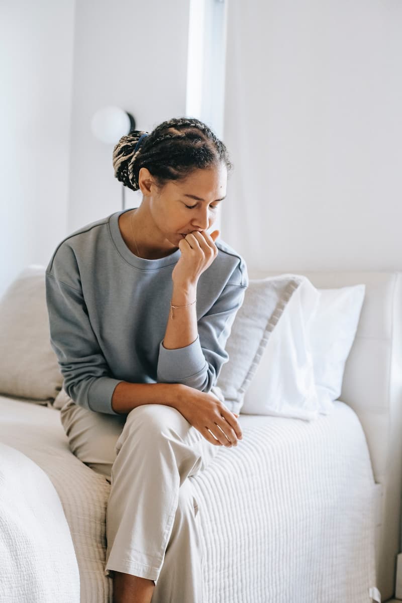 A woman sits on the edge of a bed with her head resting in one hand with a sad expression on her face.